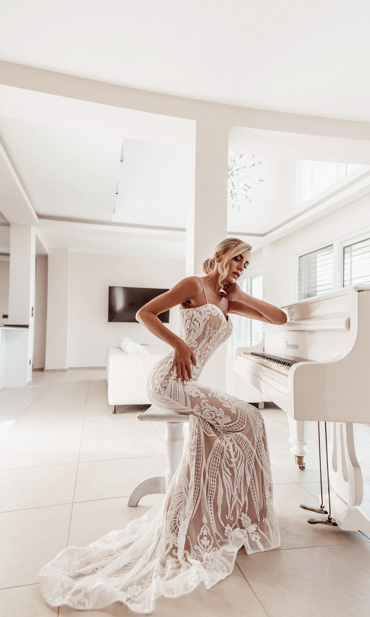 A woman in the White Lace Bodycon Bridal Gown: Tina Holly BA999 sits at a white grand piano in a bright, modern room with large windows.